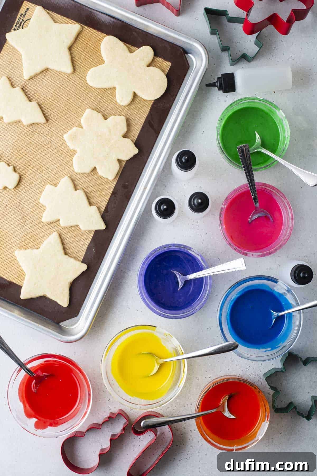 Christmas shaped cookies lined in rows on a cookie sheet with bowls of colored icing with spoons beside it, empty squeeze bottles and cookie cutters.