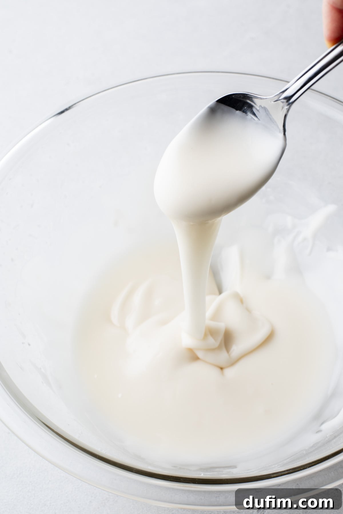 Royal icing in a clear glass bowl with a spoon full of icing above it, showing icing drizzling down into the bowl.