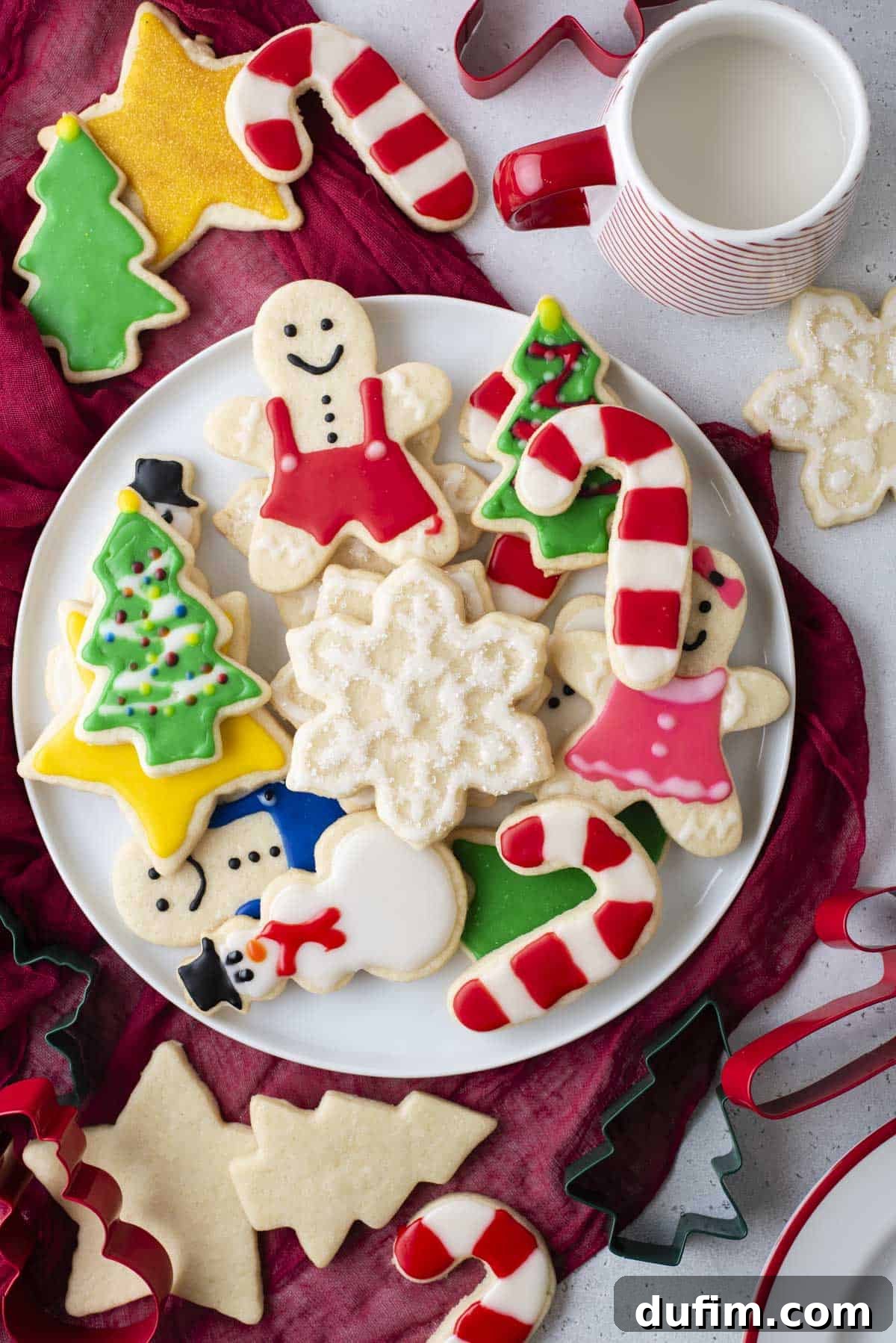 A plate full of Christmas cookies decorated with royal icing on top of a red towel, with more cookies, cookie cutters, and a cup of milk around it.