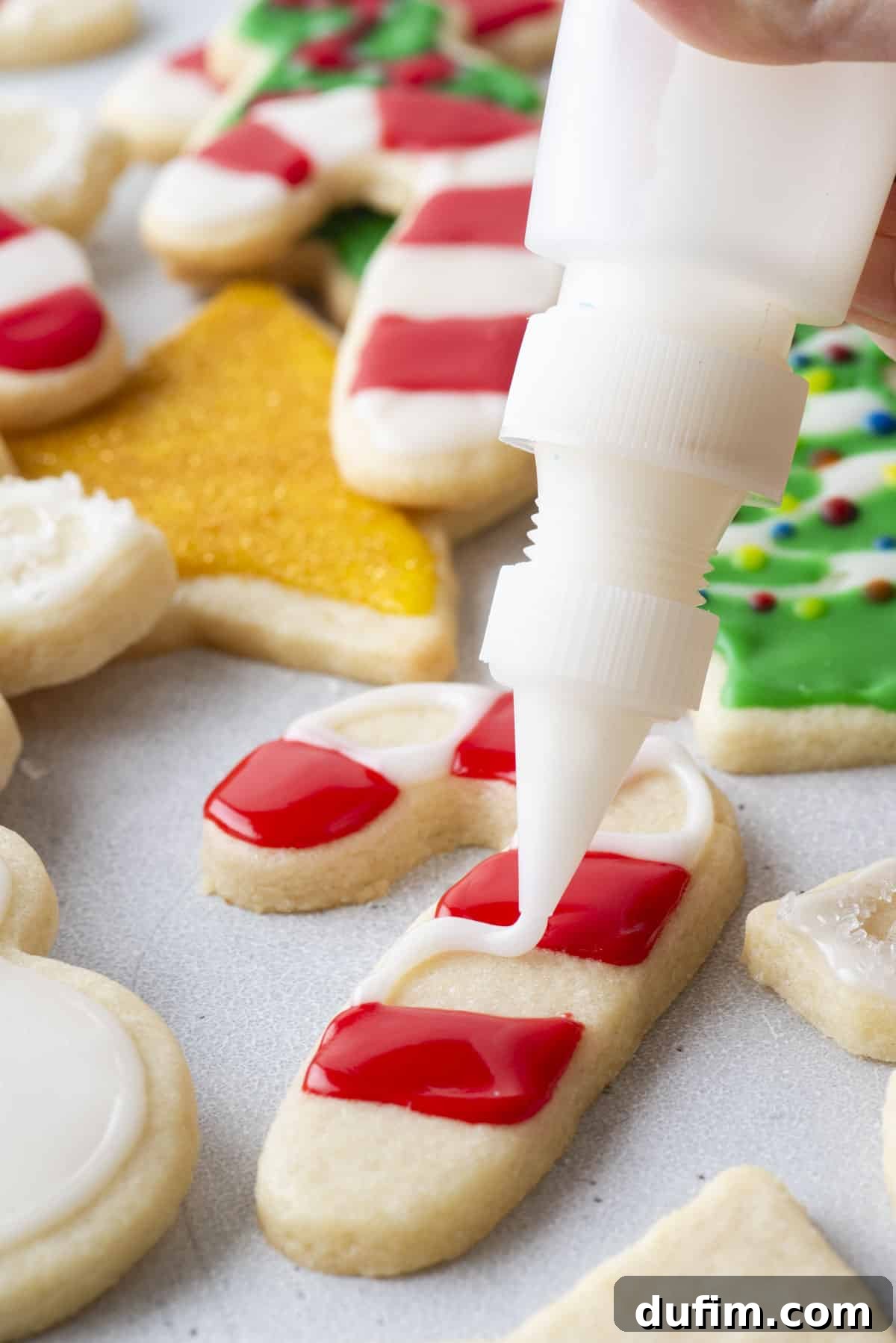 White royal icing being piped in between red icing on a candy cane-shaped cookie, with more Christmas-themed cookies in the background.