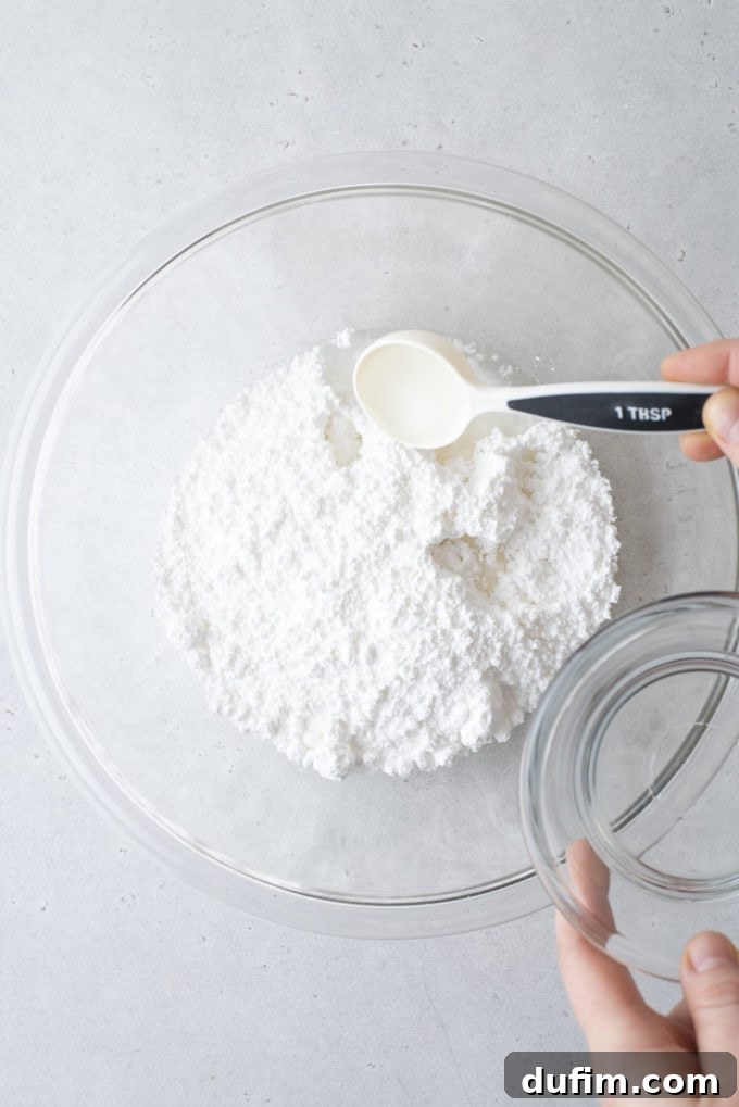 Ingredients for royal icing in a clear glass bowl with water being added using a small measuring spoon.