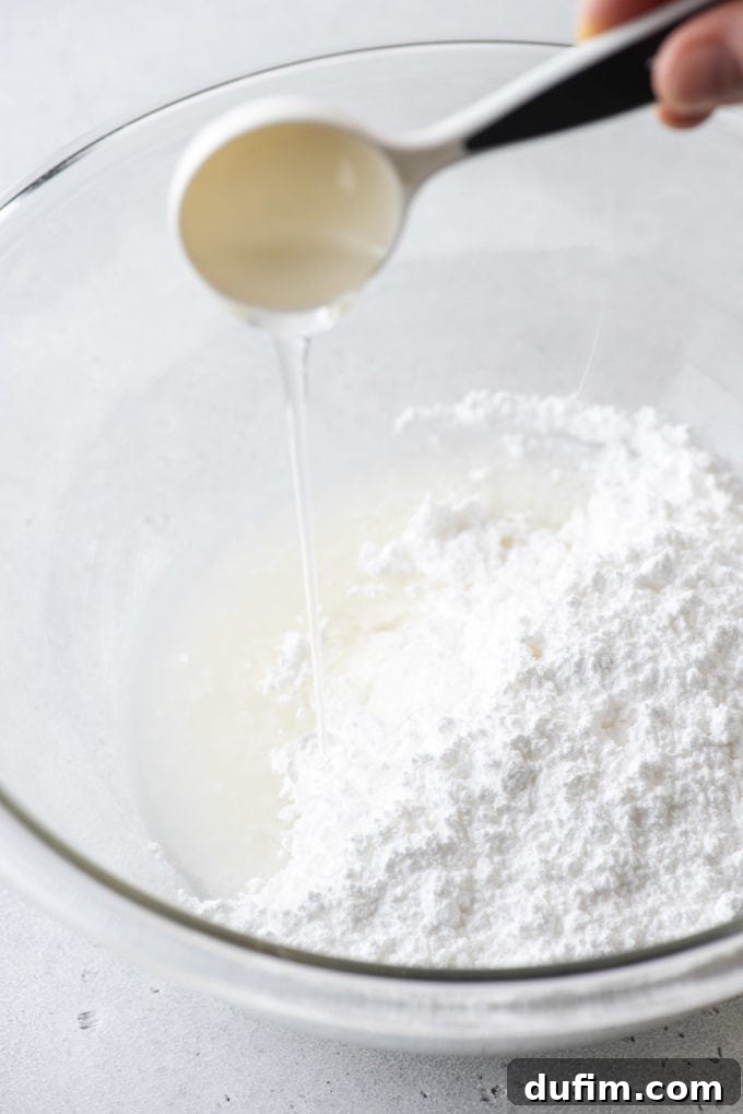 Corn syrup being added to a clear glass bowl of ingredients for royal icing.