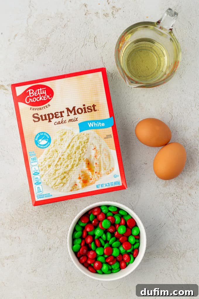 Ingredients for festive Christmas sprinkle cookies on a countertop: a box of white cake mix, vegetable oil, two eggs, and a small bowl brimming with green and white M&M's, ready for mixing.