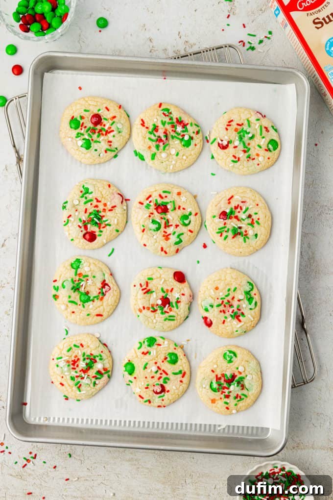 Four neat rows of freshly baked Christmas sprinkle cookies, cooling on a wire rack over a parchment-lined baking sheet. Each cookie is dotted with festive green and white sprinkles.
