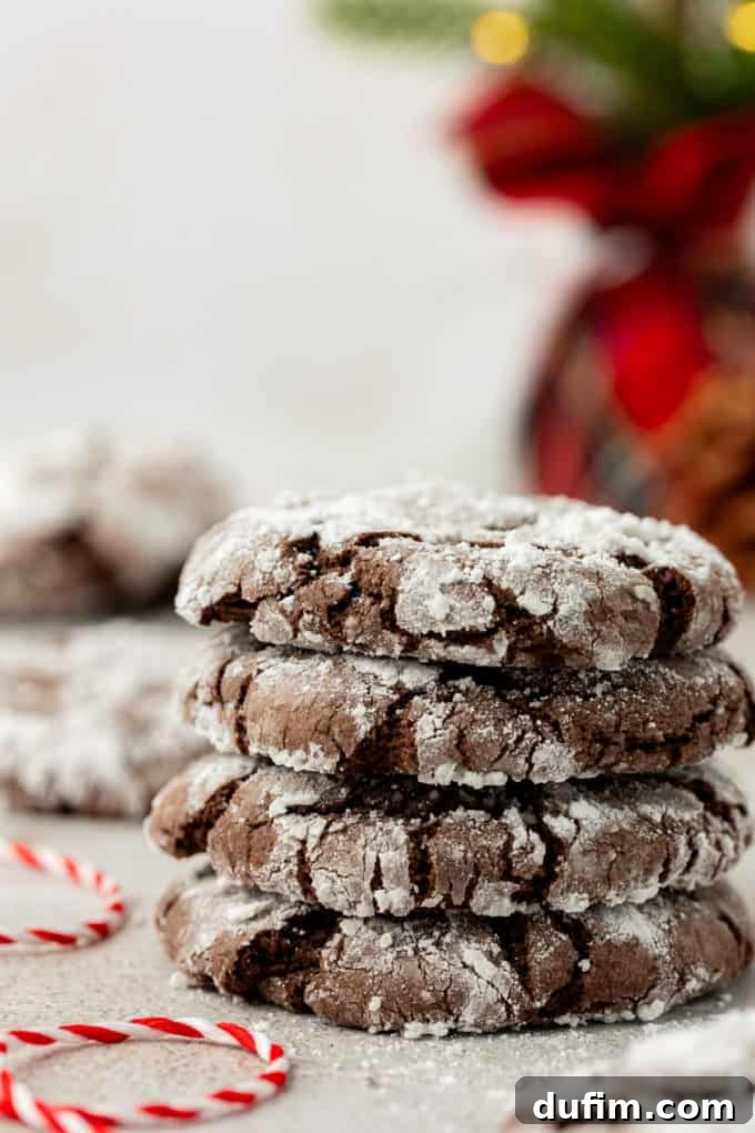 A stack of four perfectly crinkled chocolate crinkle cake mix cookies, dusted with powdered sugar, with a decorative red and white string beside them and more cookies visible in the background.
