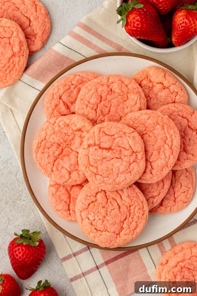 A beautiful pile of strawberry cake mix cookies on a white plate with a brown rim, resting on a tan and pink kitchen towel. More cookies, fresh strawberries, and a small white bowl of strawberries are scattered around, emphasizing the fruity theme.