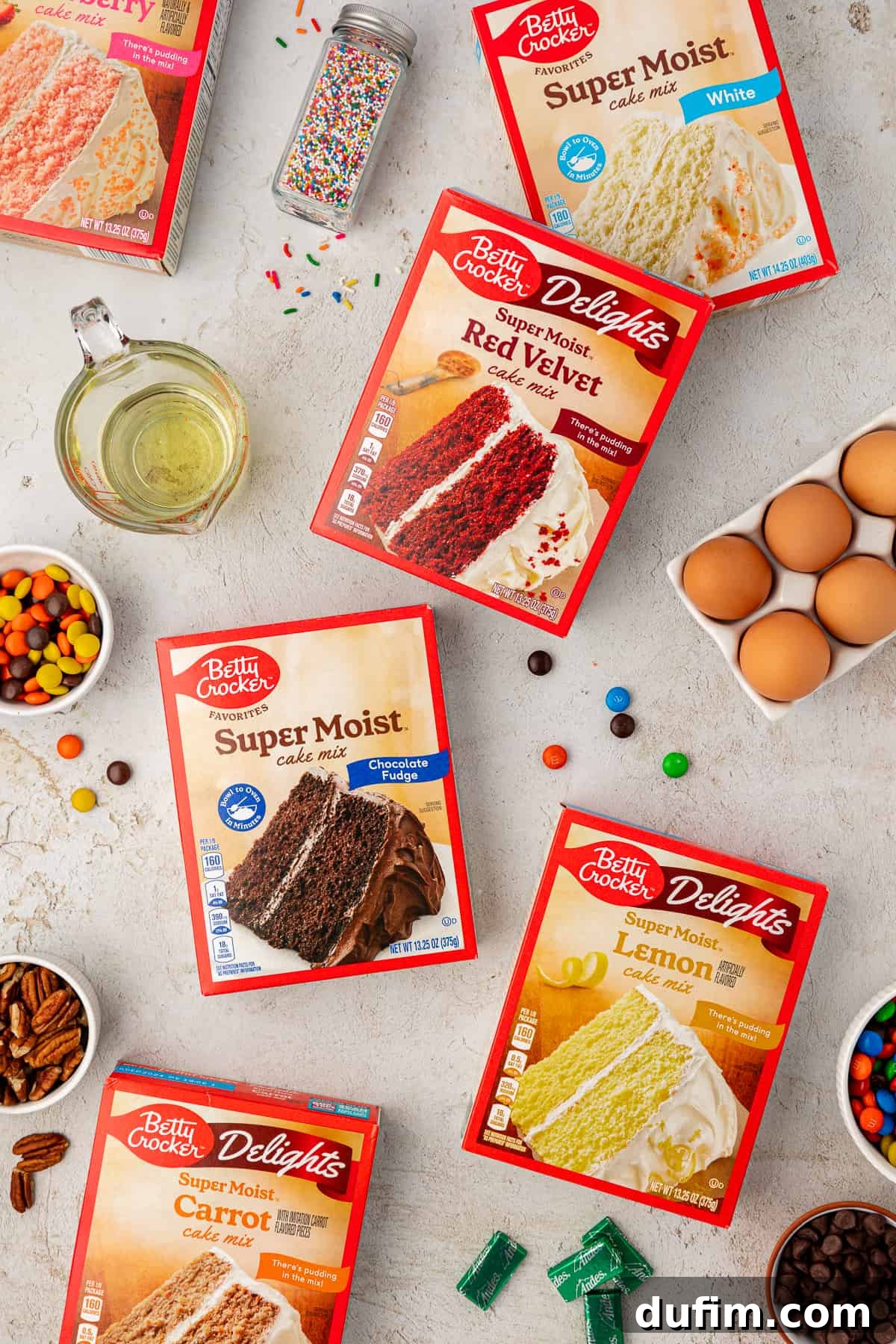 An array of essential ingredients for making diverse cake mix cookies. Featured are boxes of strawberry, white, red velvet, chocolate, and lemon cake mix, alongside vegetable oil, fresh eggs, M&M's, nuts, Reese's Pieces, colorful sprinkles, and Andes mints, highlighting the vast customization potential.