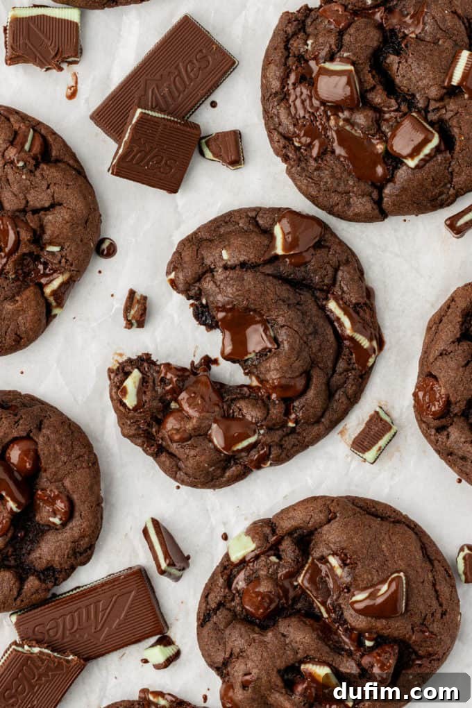 Freshly baked Andes mint cookies on white paper, showcasing their gooey melted chocolate centers and visible bits of Andes mints sprinkled throughout, with one cookie partially pulled apart.