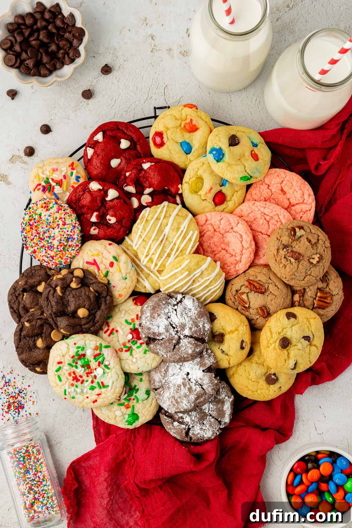 A charming circular arrangement of assorted cake mix cookies in various flavors on a wire rack, placed over a festive red towel. Colorful sprinkles, M&M's, chocolate chips, and two glasses of milk with red and white straws complete this inviting dessert scene.