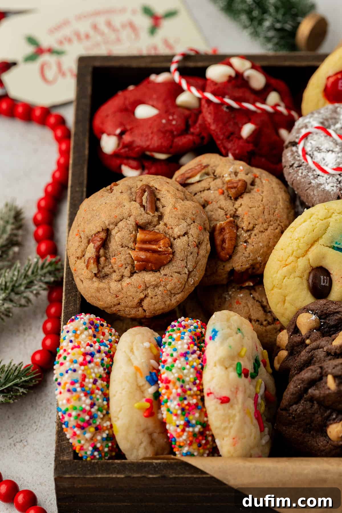A charming wooden box filled with an assortment of beautifully baked cake mix cookies in various flavors, nestled on brown paper. The scene is adorned with festive red beads, fragrant pine tree stems, and red and white string, perfect for a cozy holiday gift.