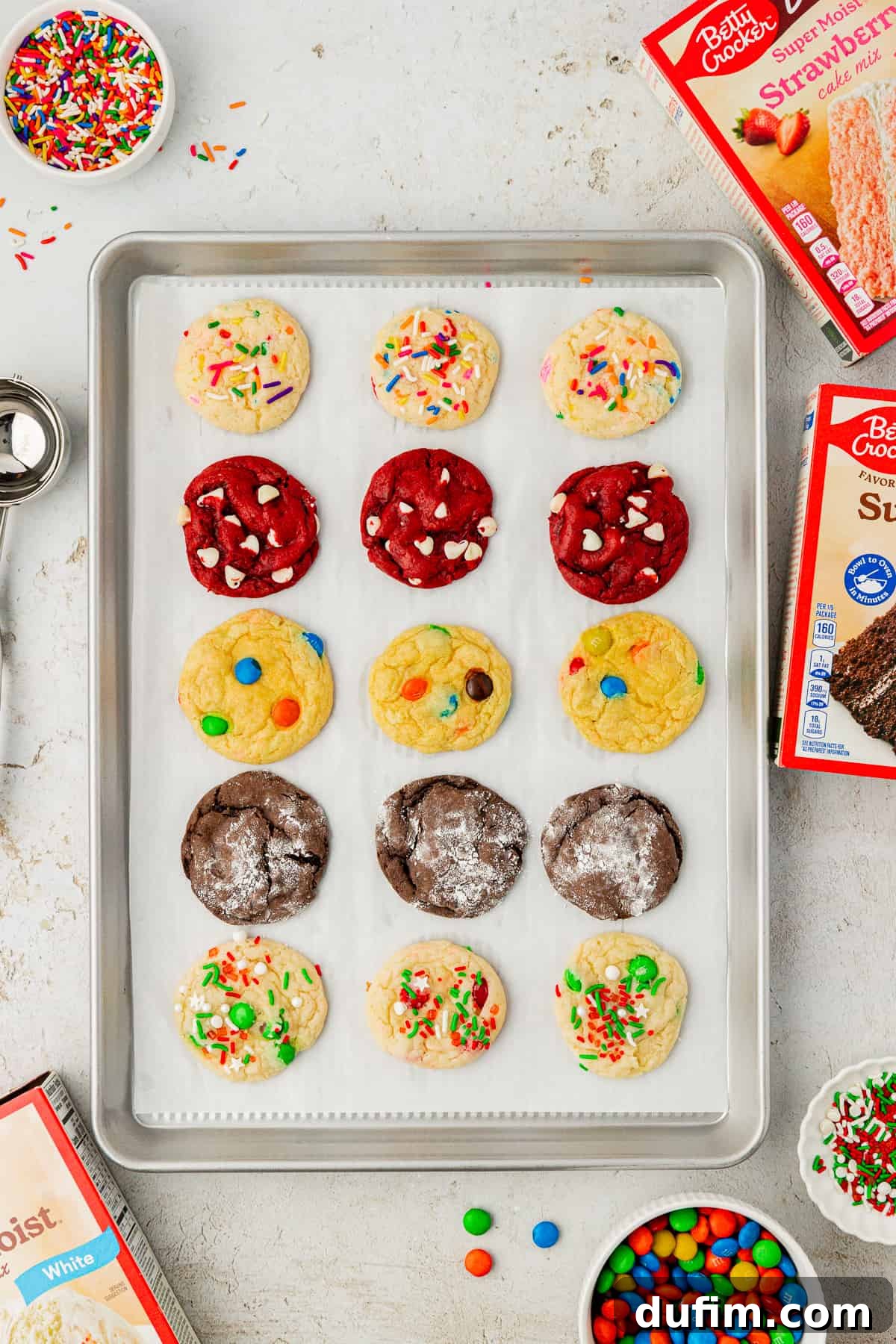 An organized display of various cake mix cookie flavors, neatly arranged in rows on a parchment-lined baking sheet. Surrounding the cookies are colorful boxes of cake mix, vibrant sprinkles, M&M's, and a handy cookie scoop, highlighting the ease and variety of this recipe.