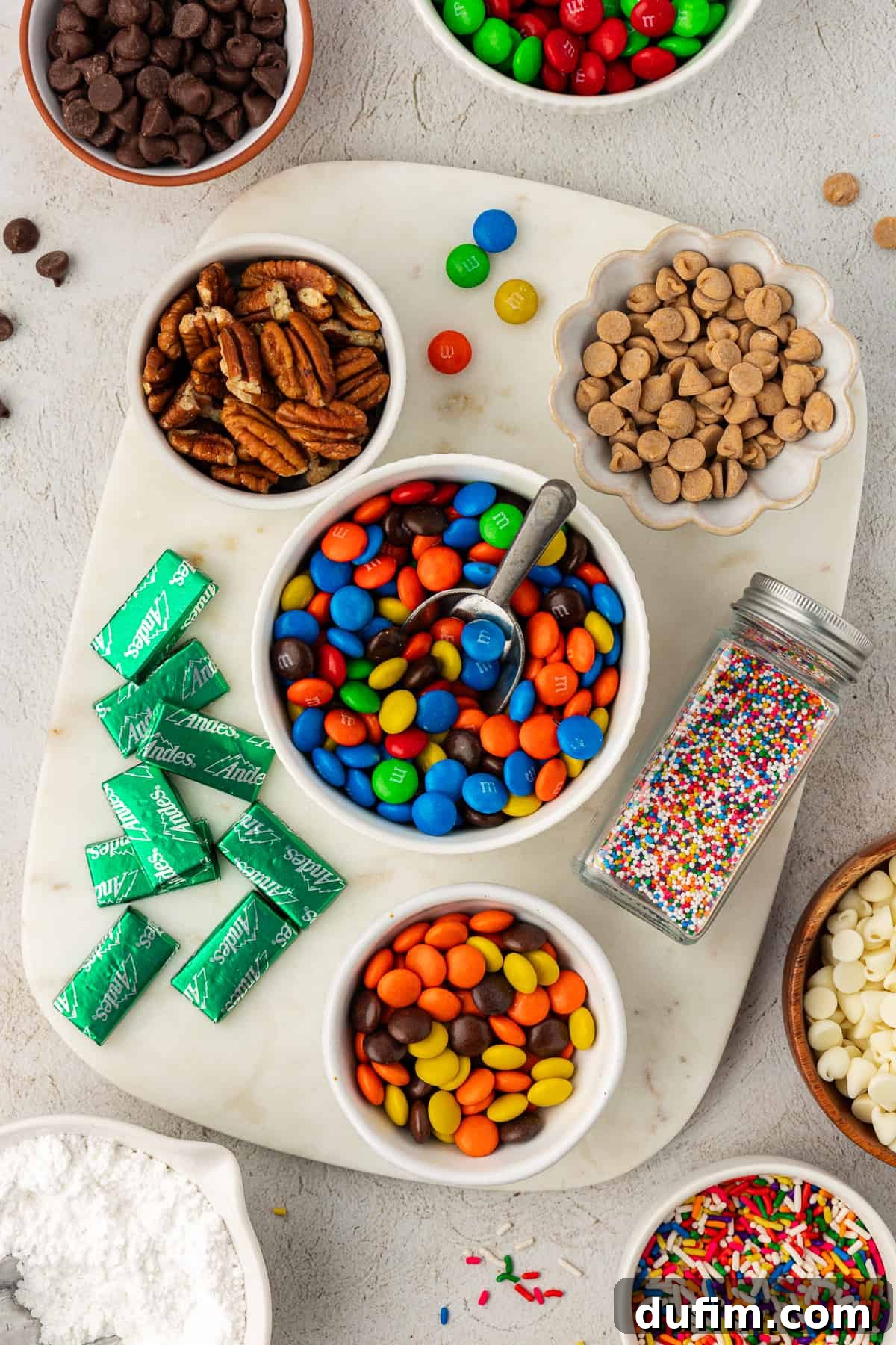A vibrant assortment of popular mix-ins for cake mix cookies, including classic chocolate chips, colorful M&M's, crunchy pecans, creamy peanut butter chips, festive red & green M&M's, cool Andes mints, bright sprinkles, luxurious white chocolate chips, and delicate powdered sugar for dusting.