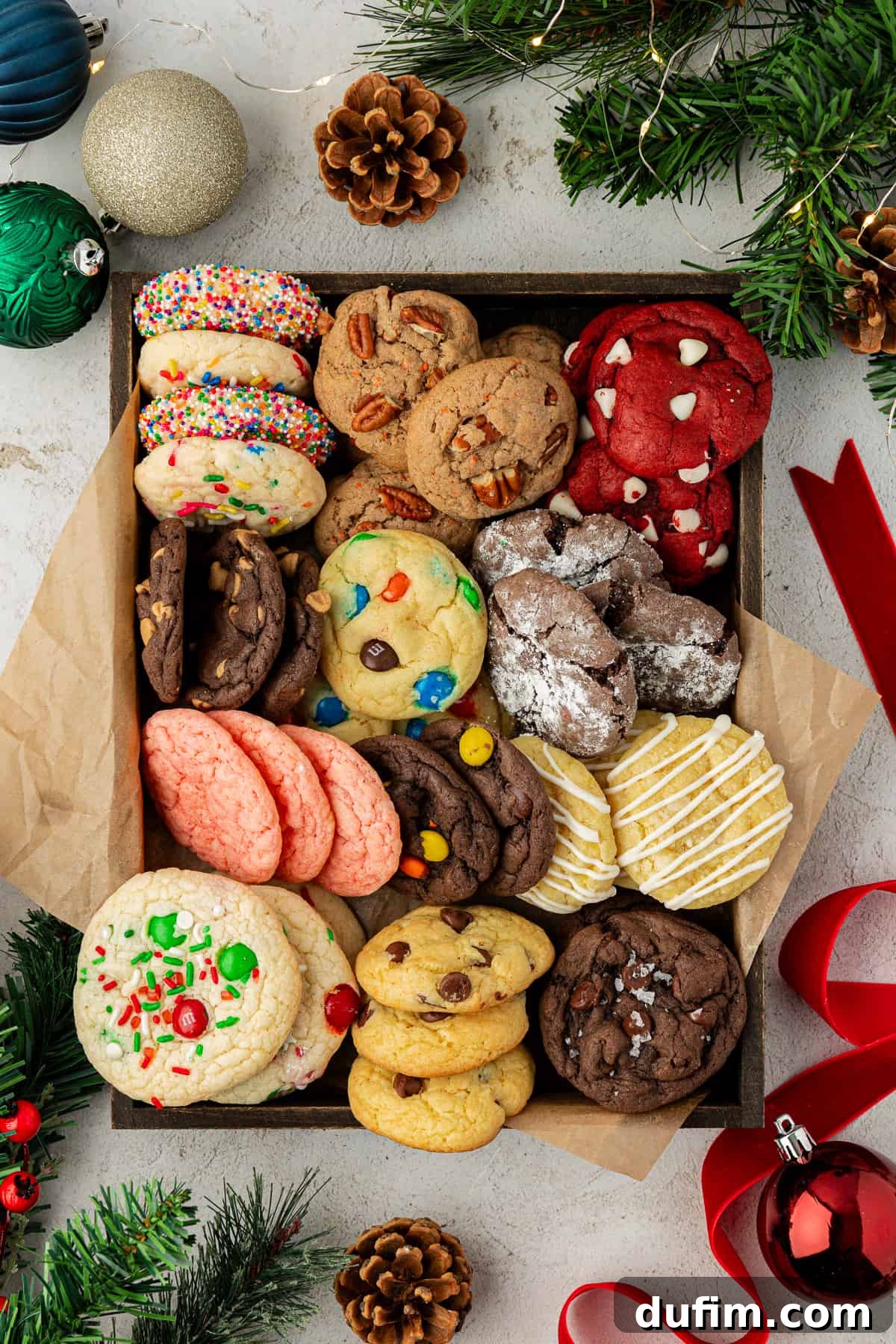 A festive box filled with an assortment of cake mix cookie flavors, beautifully arranged on brown paper and surrounded by Christmas ornaments, red ribbon, pine cones, and evergreen branches, creating a perfect holiday gift presentation.