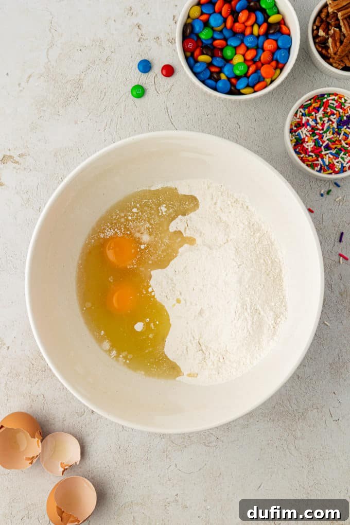 A white bowl holds the dry cake mix and cracked eggs, surrounded by empty eggshells, a colorful bowl of sprinkles, a vibrant bowl of M&M's, and a bowl of nuts, illustrating the initial mixing phase for cake mix cookies.