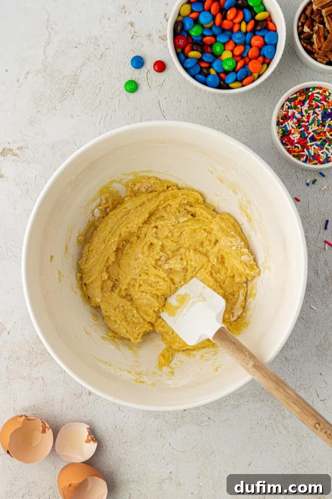 Thick, slightly sticky cake mix cookie dough in a white mixing bowl, being combined with a white and wooden spatula. Empty eggshells, bowls of sprinkles, M&M's, and nuts are artfully arranged around the bowl, indicating the next steps.