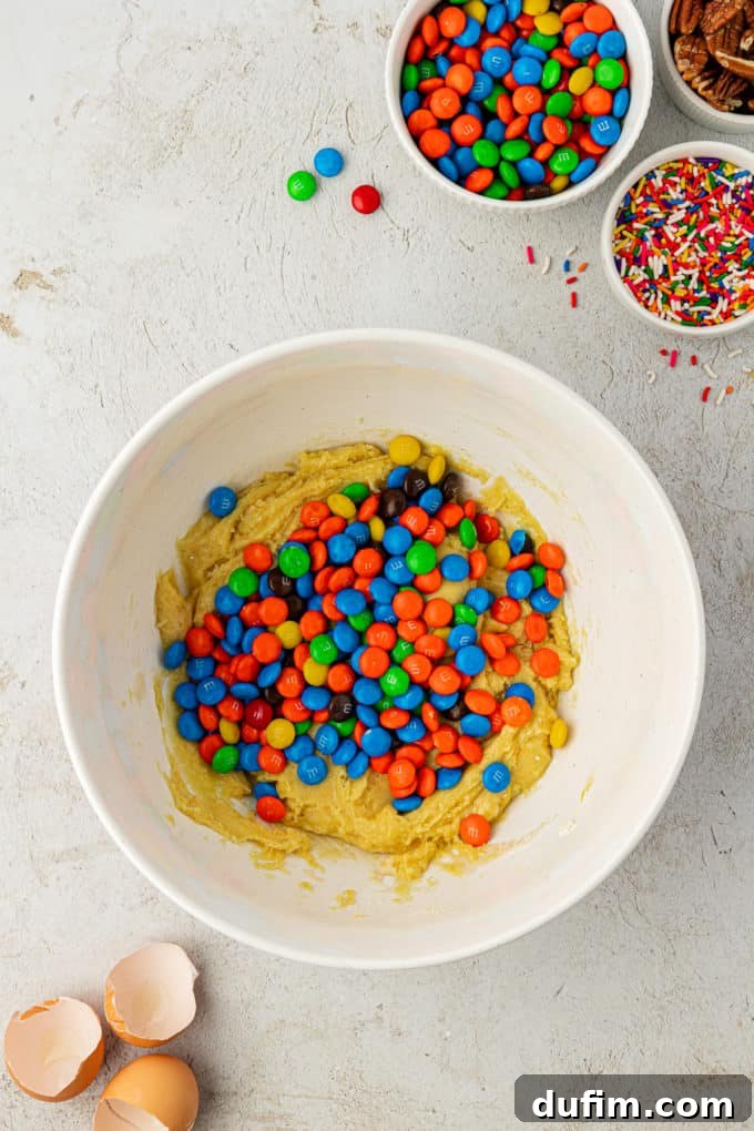 Cake mix cookie batter in a white bowl, generously topped with a pile of colorful M&M's, poised for mixing. Surrounding elements like eggshells, sprinkles, and nuts suggest the full baking process.
