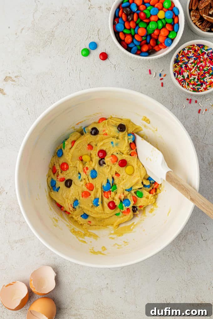 Vibrant M&M's are perfectly mixed into soft cake mix cookie batter within a white bowl, actively stirred by a white and wooden spatula. Bowls of sprinkles, M&M's, and nuts remain nearby, indicating a creative baking session.