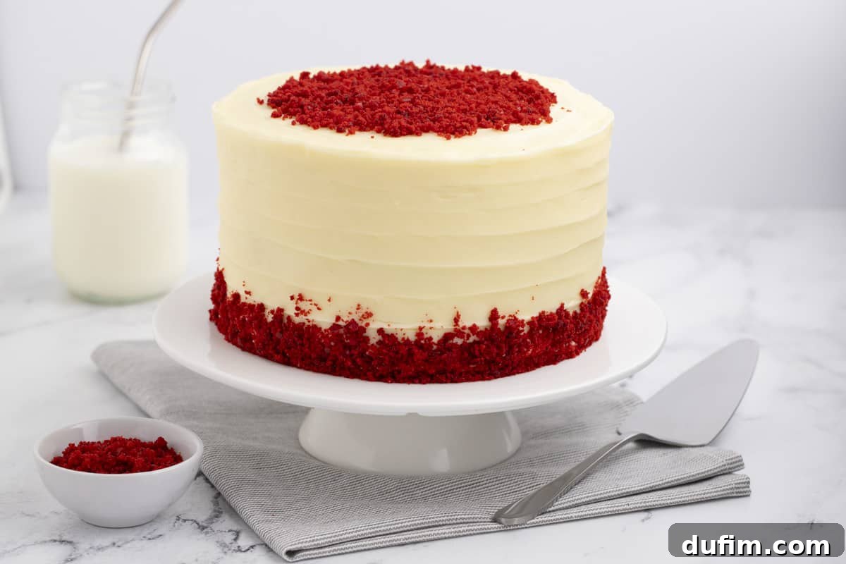 Homemade Red Velvet Cake Perfection 2 A beautifully frosted red velvet cake with cream cheese frosting, elegantly presented on a white cake stand. Beside it, a bowl of cake crumbs, a serving knife, and a refreshing glass of milk complete the inviting scene.