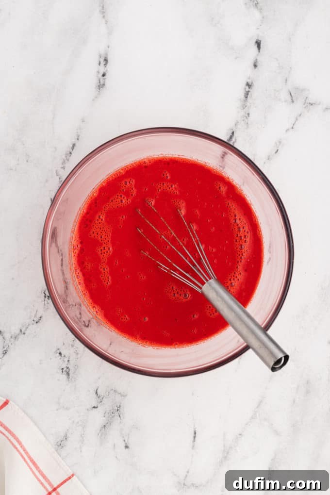 Homemade Red Velvet Cake Perfection 11 The vibrant red wet ingredients for red velvet cake are whisked in a clear glass bowl, creating a rich, uniform color and smooth consistency.