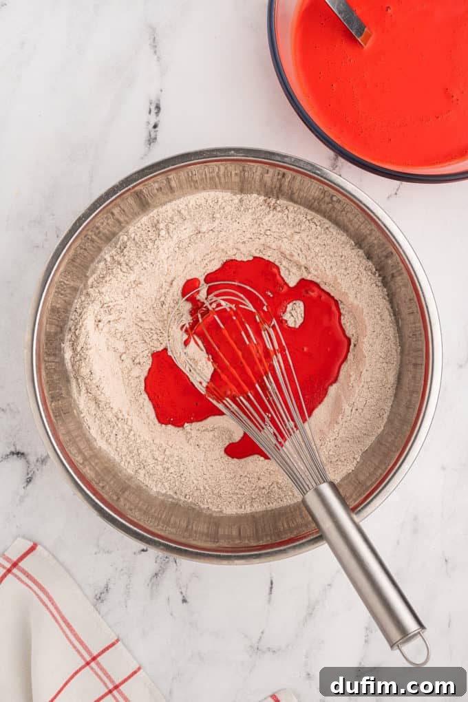 Homemade Red Velvet Cake Perfection 12 The bright red wet ingredients for red velvet cake are poured into a silver bowl containing the dry ingredients and gently mixed with a wire whisk, ensuring an even, vibrant batter.
