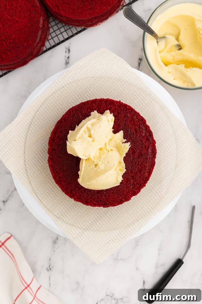 Homemade Red Velvet Cake Perfection 16 The initial layer of red velvet cake is carefully placed on a cake stand, topped with a generous dollop of cream cheese frosting. A bowl of additional frosting and a rack of cake layers are in the background.