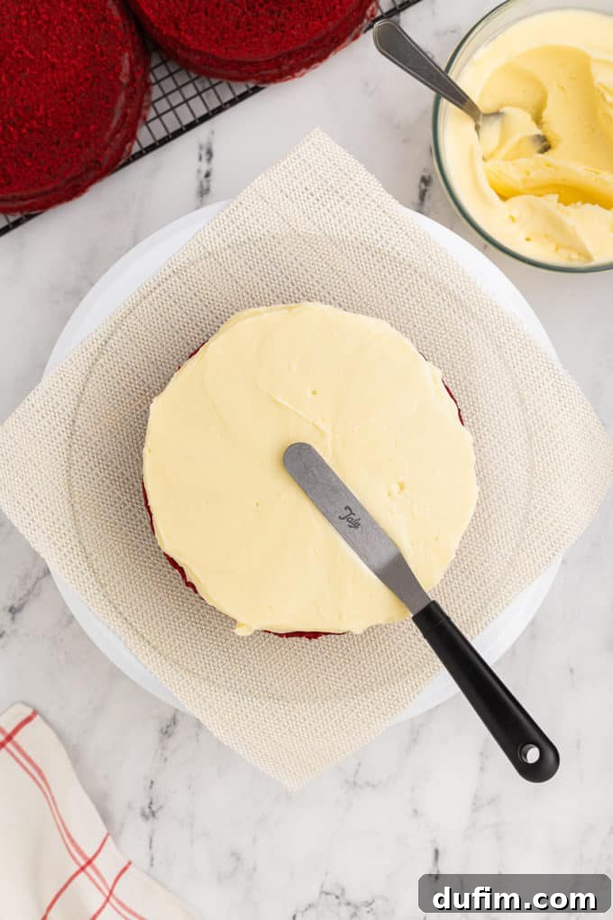 Homemade Red Velvet Cake Perfection 17 A red velvet cake layer on a cake stand is being beautifully frosted with cream cheese frosting. A bowl of extra frosting and cooled cake layers on a wire rack are in the background.