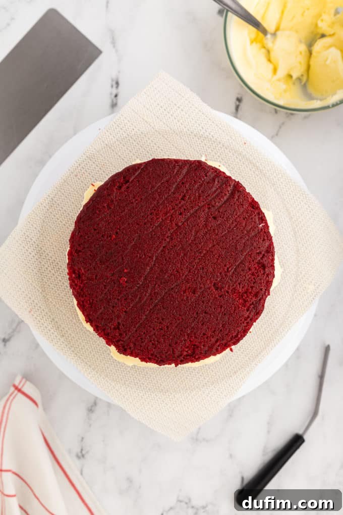 Homemade Red Velvet Cake Perfection 18 A layered red velvet cake sits proudly on a cake stand, awaiting its next layer of frosting. A bowl of creamy frosting and additional cake layers on a wire rack are visible nearby.