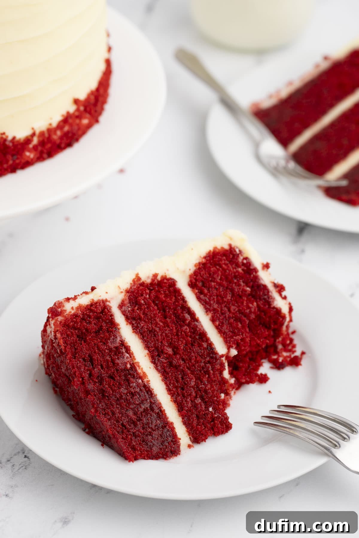 Homemade Red Velvet Cake Perfection 3 A perfect slice of layered red velvet cake, complete with generous cream cheese frosting, rests on a white plate with a fork. In the background, another slice and the main cake hint at its irresistible appeal.