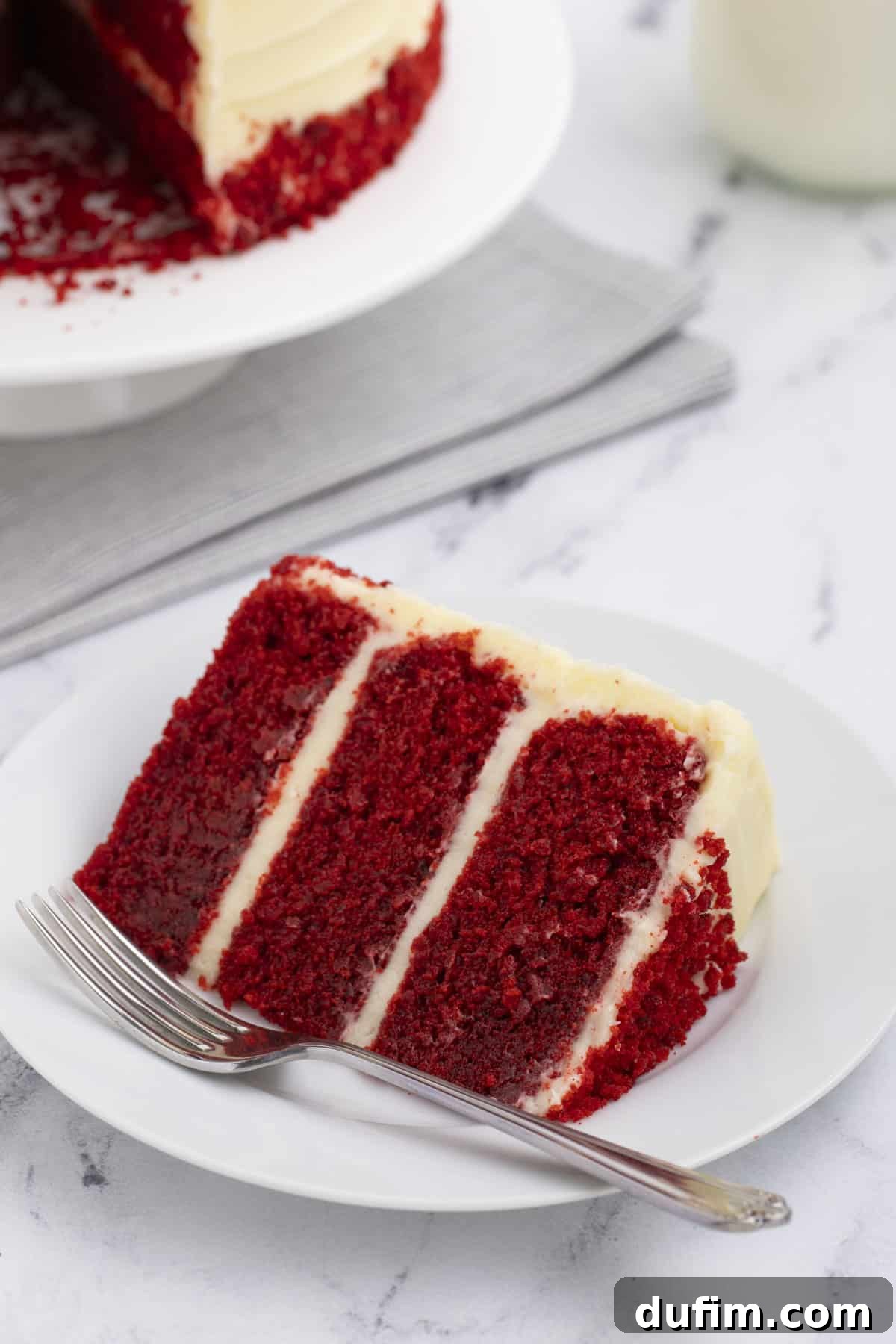 Homemade Red Velvet Cake Perfection 21 A delicate slice of red velvet cake, adorned with fluffy cream cheese frosting, rests on a small white plate with a fork. The rest of the cake is visible in the background, set on a larger white plate atop a cozy grey towel.