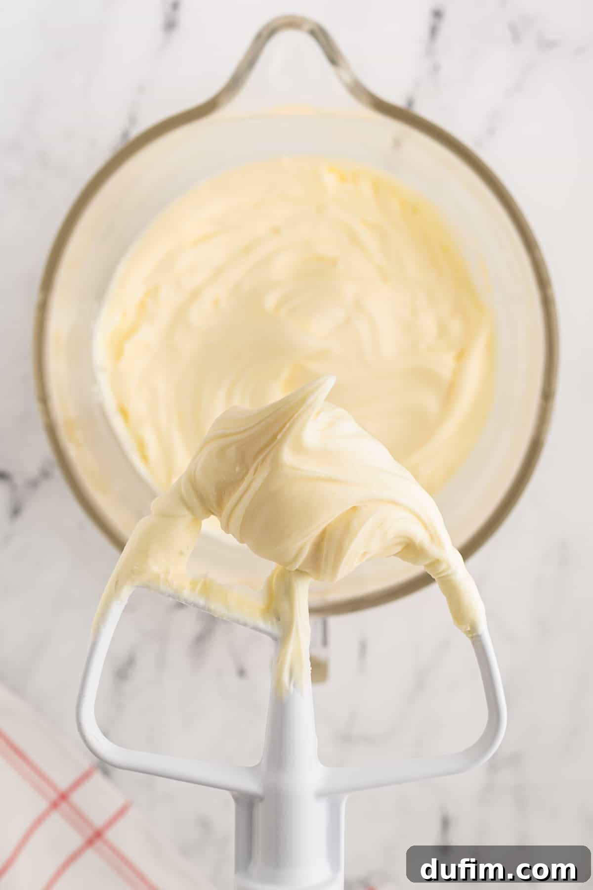 Homemade Red Velvet Cake Perfection 22 A close-up of creamy cream cheese frosting in a mixing bowl, with the mixer's whisk attachment lifted above it, generously coated in the decadent frosting.