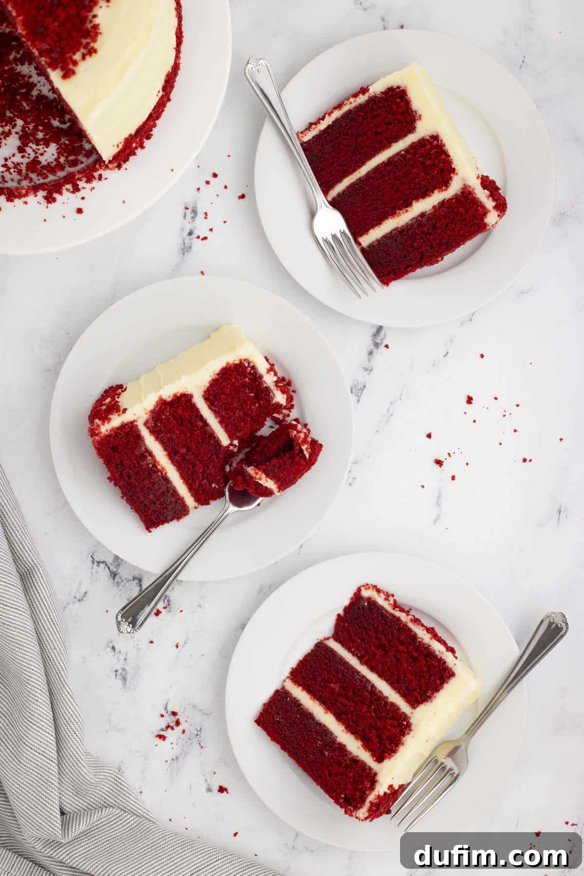 Homemade Red Velvet Cake Perfection 23 An inviting overhead shot showcasing three perfectly sliced red velvet cake portions on white plates with forks, artfully arranged around a larger white plate holding the remaining cake, highlighting its vibrant color and luscious frosting.