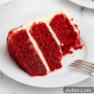 A slice of layered red velvet cake with cream cheese frosting on a white plate with a fork leaning on it