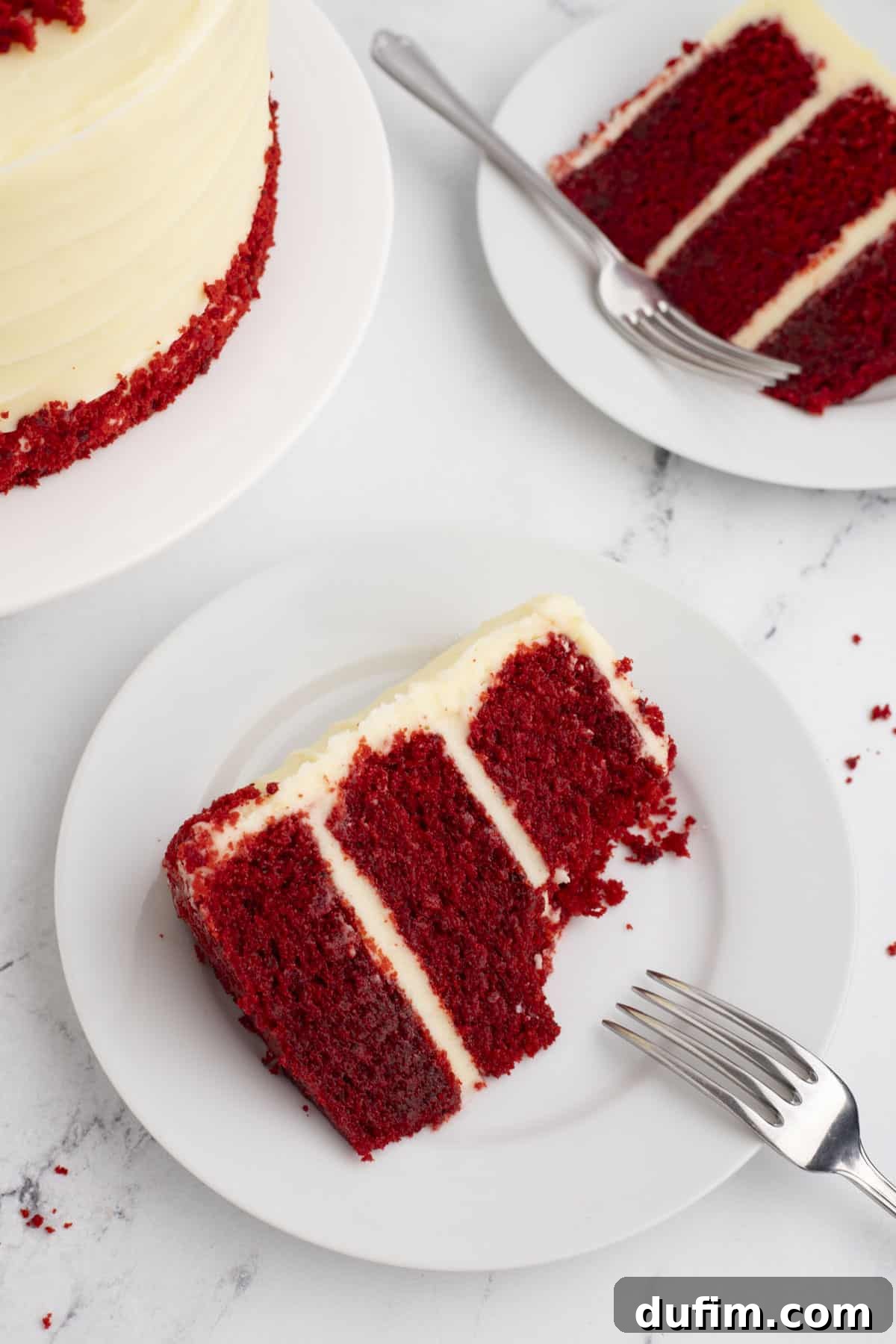 Homemade Red Velvet Cake Perfection 6 A delectable slice of layered red velvet cake with a generous helping of cream cheese frosting sits on a white plate with a fork. In the soft-focus background, the remaining cake and another slice await enjoyment.