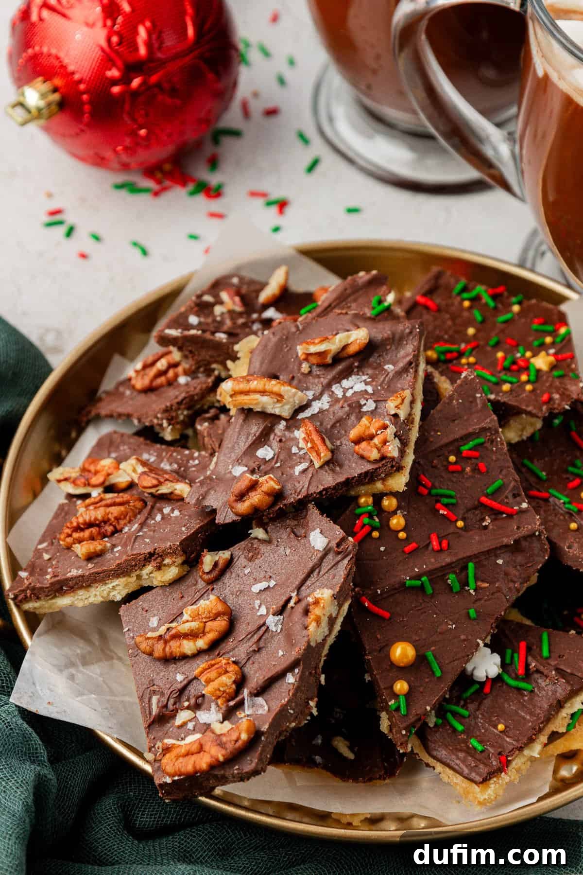 A gold plate showcasing Christmas Crack, half topped with pecans and half with colorful sprinkles, set on a white surface alongside festive decorations like a green towel, red ornament, and holiday sprinkles.