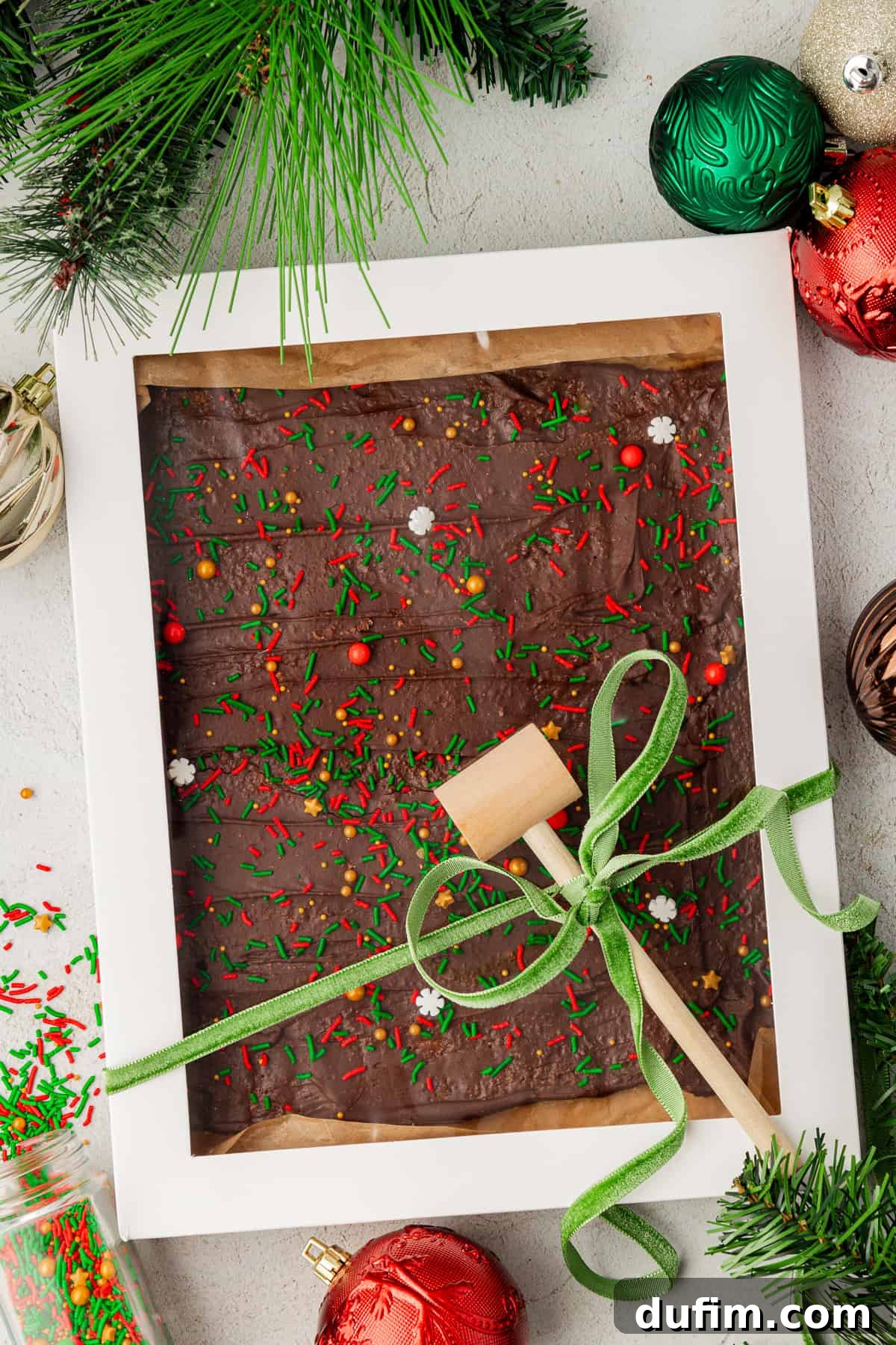 A large white gift box with a clear top, overflowing with Christmas Crack candy, tied with a green ribbon and a small wooden mallet. Festive decor surrounds it.