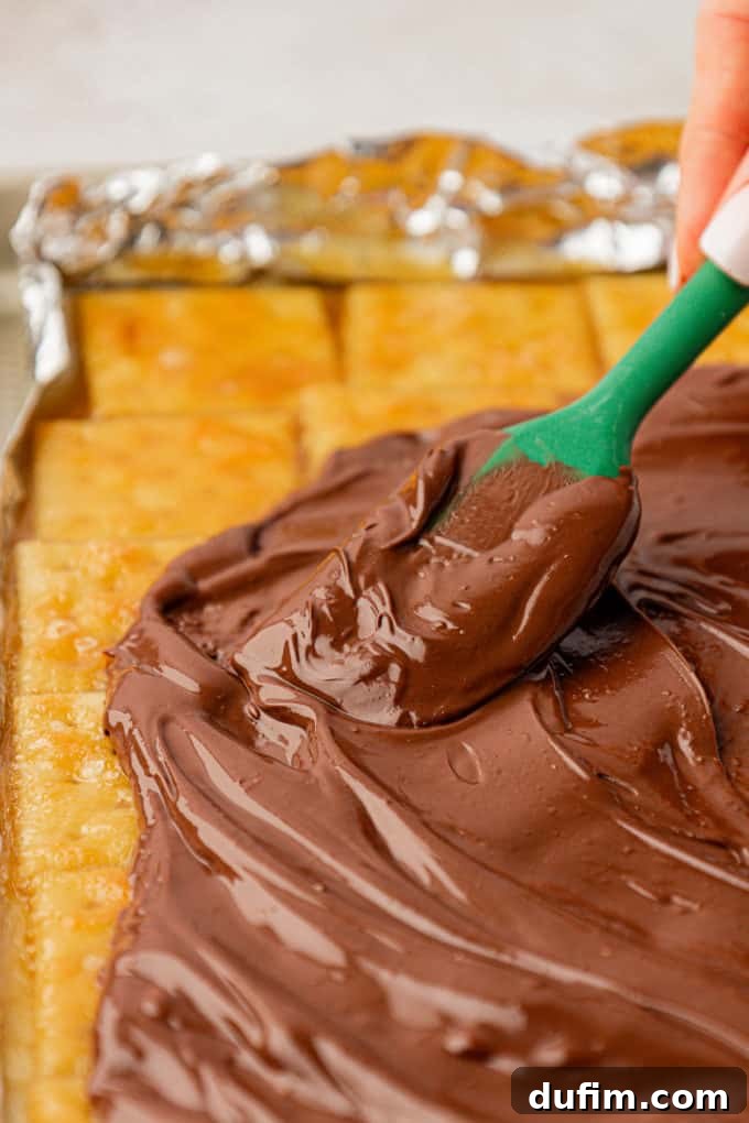 Melted chocolate being smoothly spread with a green spatula over the warm, toffee-coated saltine crackers on a baking sheet.