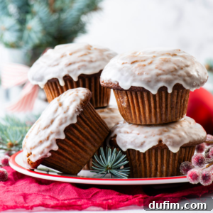 Four gingerbread muffins with white icing are stacked on a red and white plate, surrounded by festive pine branches and berries, with a red cloth underneath, creating a cozy holiday or winter theme.