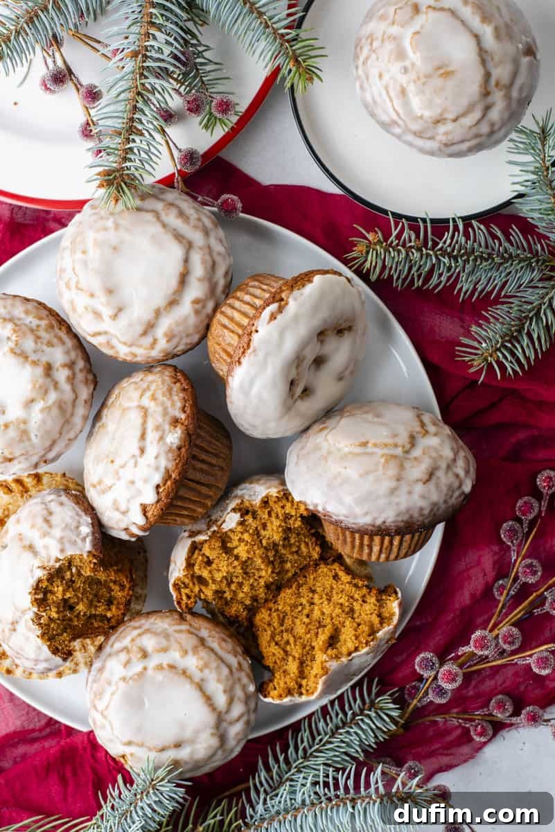 A close-up shot of several gingerbread muffins, elegantly adorned with a vanilla glaze, resting on a white plate with complementary red sugared berries and evergreen foliage.