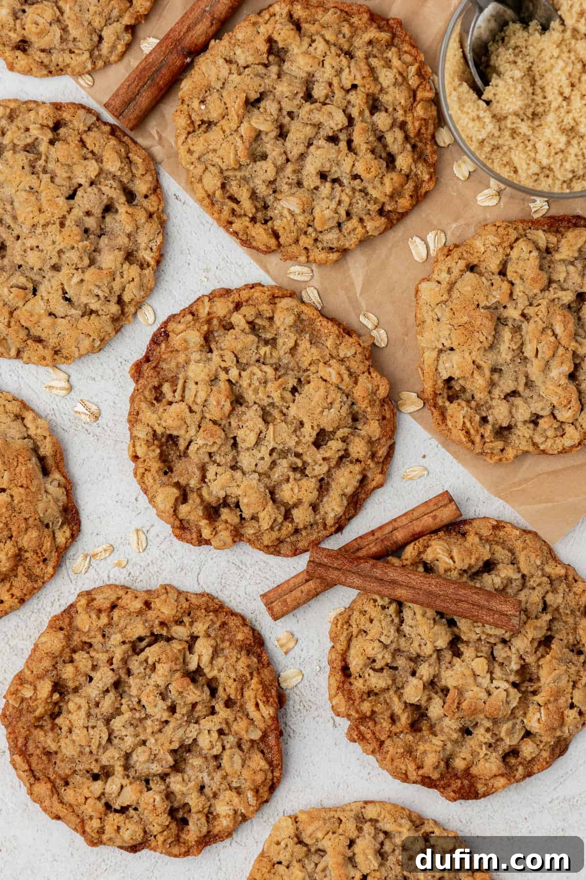 overhead view of oatmeal cookies in one layer with parchment paper, oats sprinkled around, cinnamon sticks and a cup of brown sugar