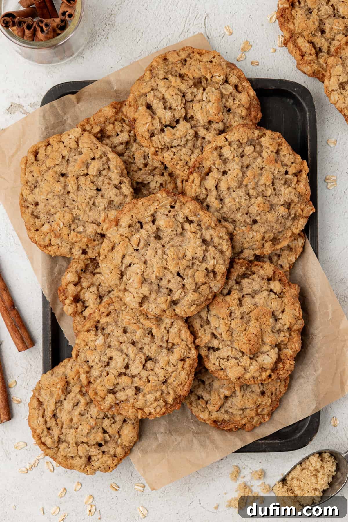 a sheet pan with parchment paper and a pile of oatmeal cookies on top with cinnamon sticks, more cookies and a spoon of brown sugar around it