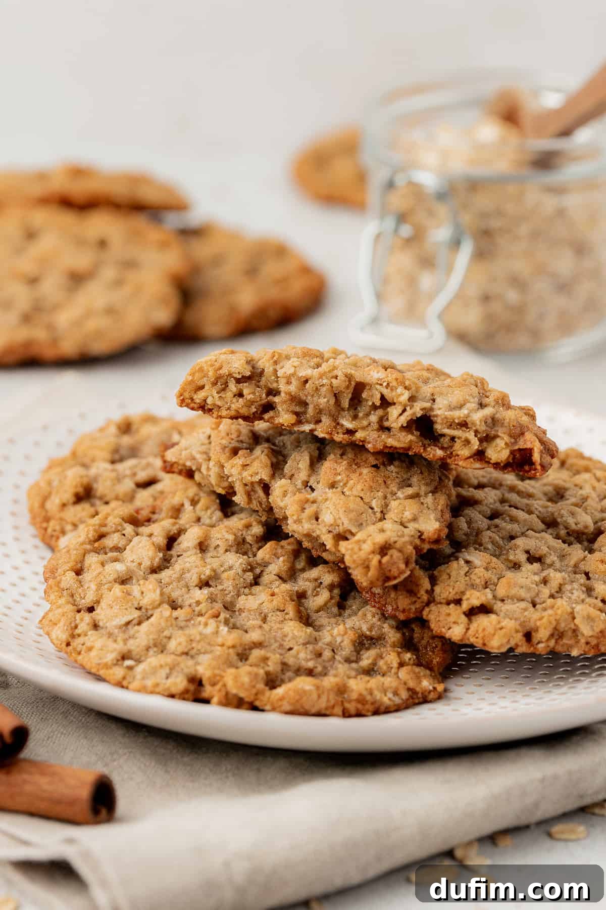 a white plate with a pile of oatmeal cookies on it on top of a tan kitchen towel with cinnamon sticks beside it, more cookies and a jar of brown sugar in the background