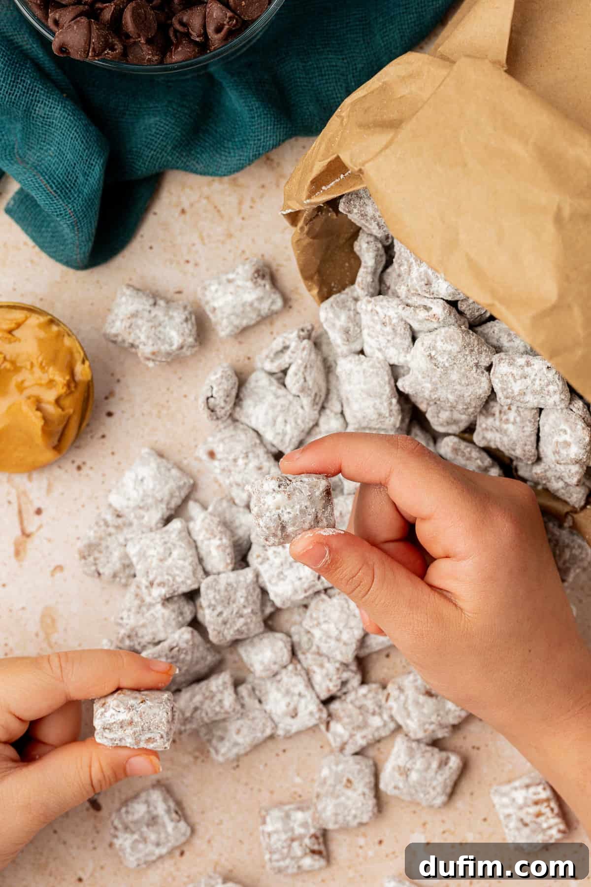 A brown bag full of puppy chow laying on its side with puppy chow spilled out, two hands holding pieces of puppy chow, a scoop of peanut butter beside it and a dark teal towel