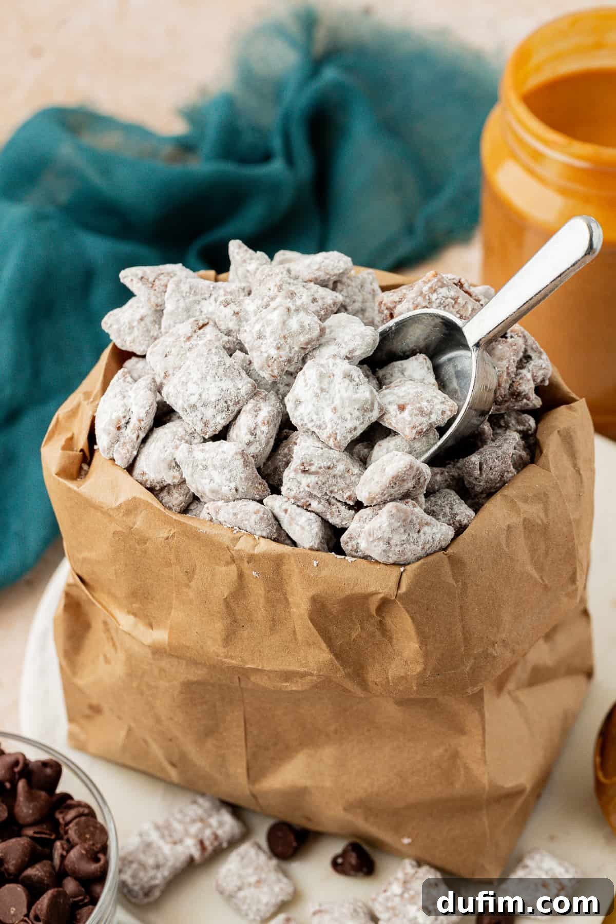 a brown bag full of puppy chow with a silver measuring spoon in it, and a jar of peanut butter and dark teal towel in the background and bowl of chocolate chips beside it, with more puppy chow and chocolate chips sprinkled around