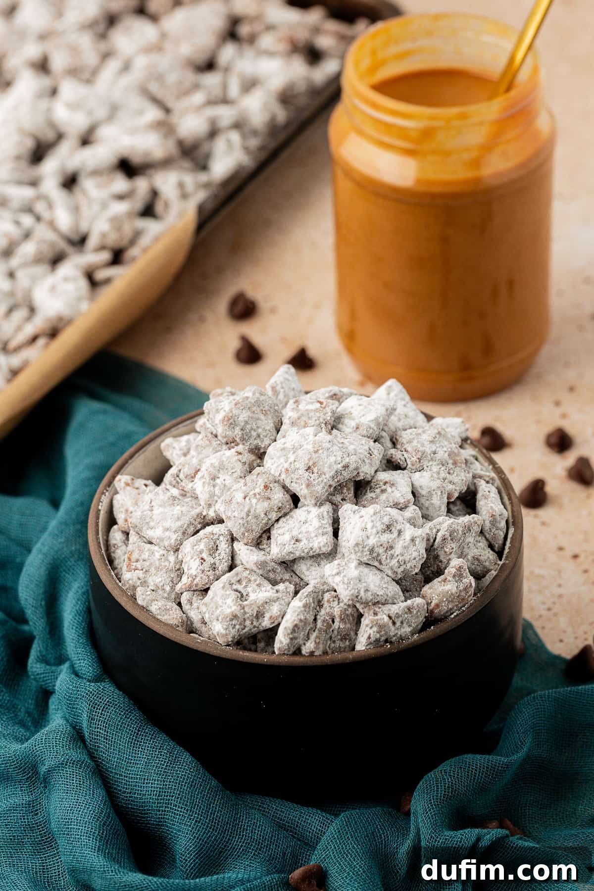 A black bowl full of puppy chow on a dark teal towel with a jar of peanut butter beside it and a sheet pan full of more puppy chow, with chocolate chips sprinkled around