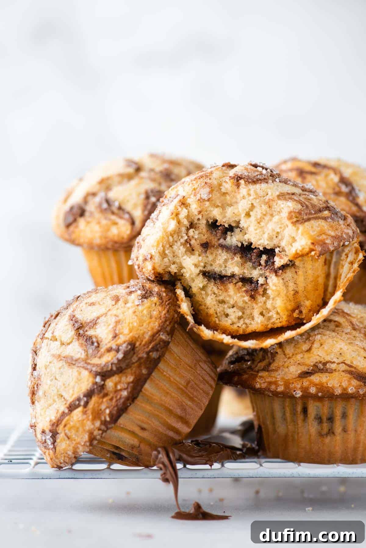 A pile of Nutella swirl muffins on a wire rack, with one muffin on its side showing the rich Nutella layers inside. A butter knife with Nutella dripping rests beside the rack.