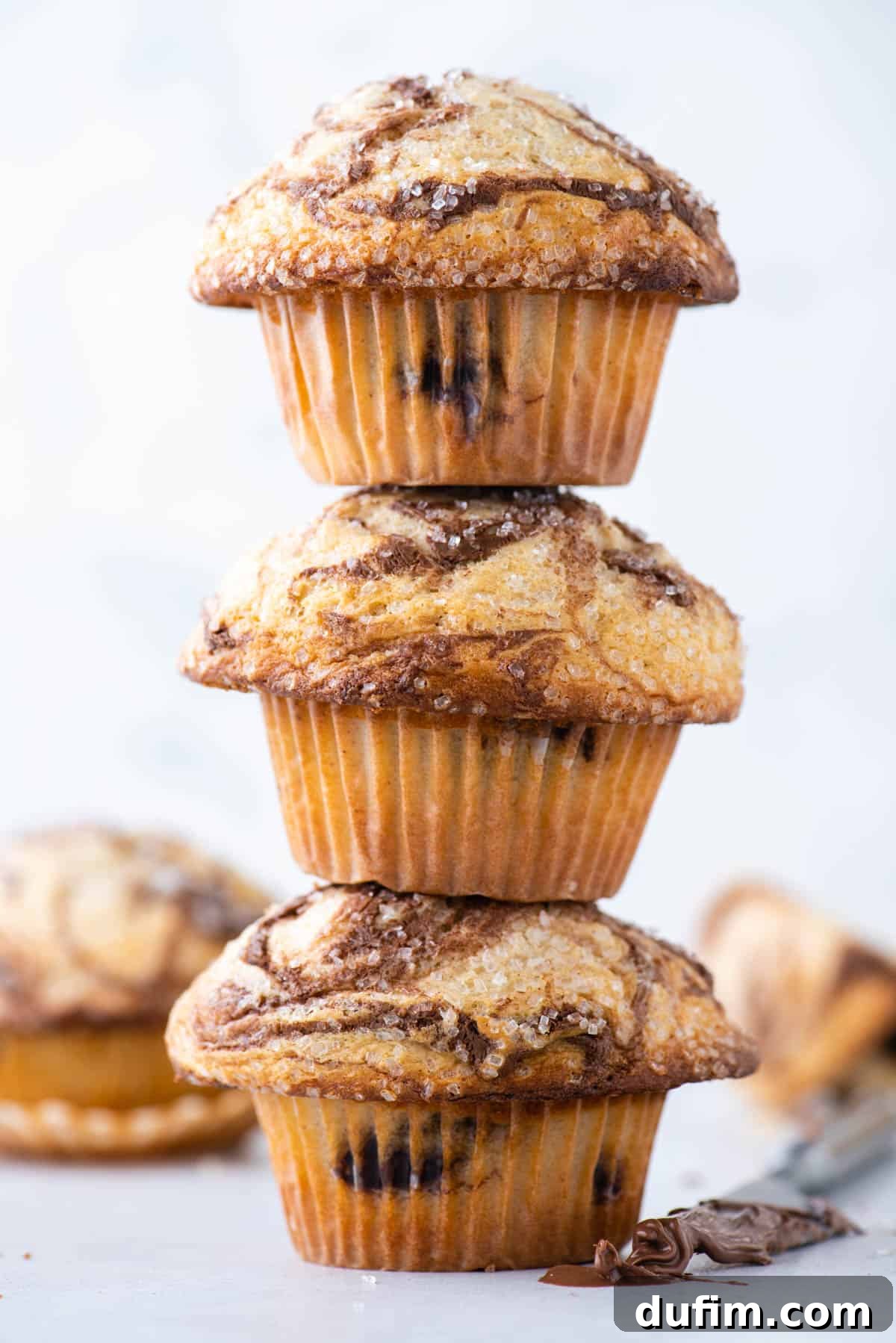 A charming tower of three Nutella swirl muffins, artfully arranged with more muffins blurred in the background. A butter knife, generously coated with Nutella, rests invitingly beside the stack.