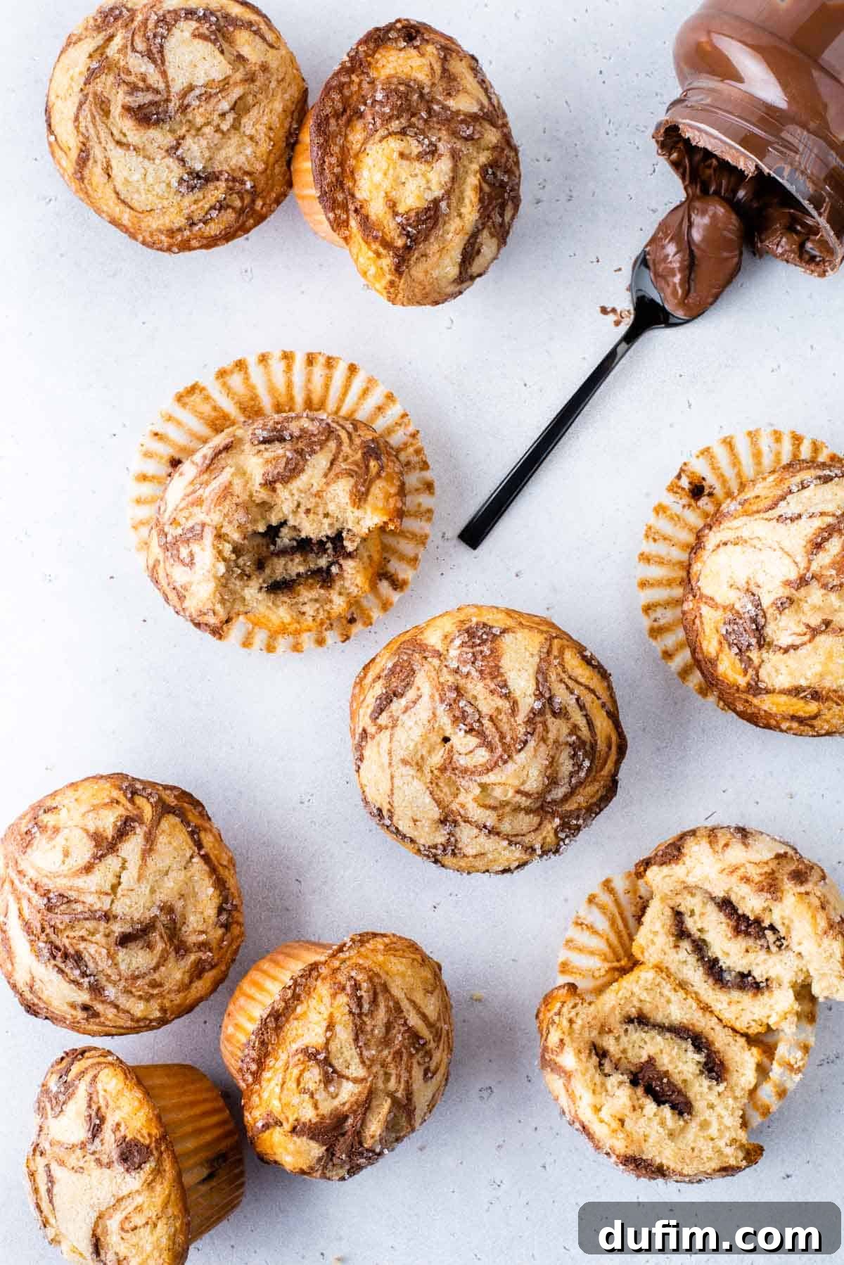 Several Nutella swirl muffins arranged on a white surface. Some are upright, some on their sides, one is cut in half revealing the exquisite Nutella layers within, and another has a bite taken out. A Nutella jar lies beside a spoon laden with the creamy spread.