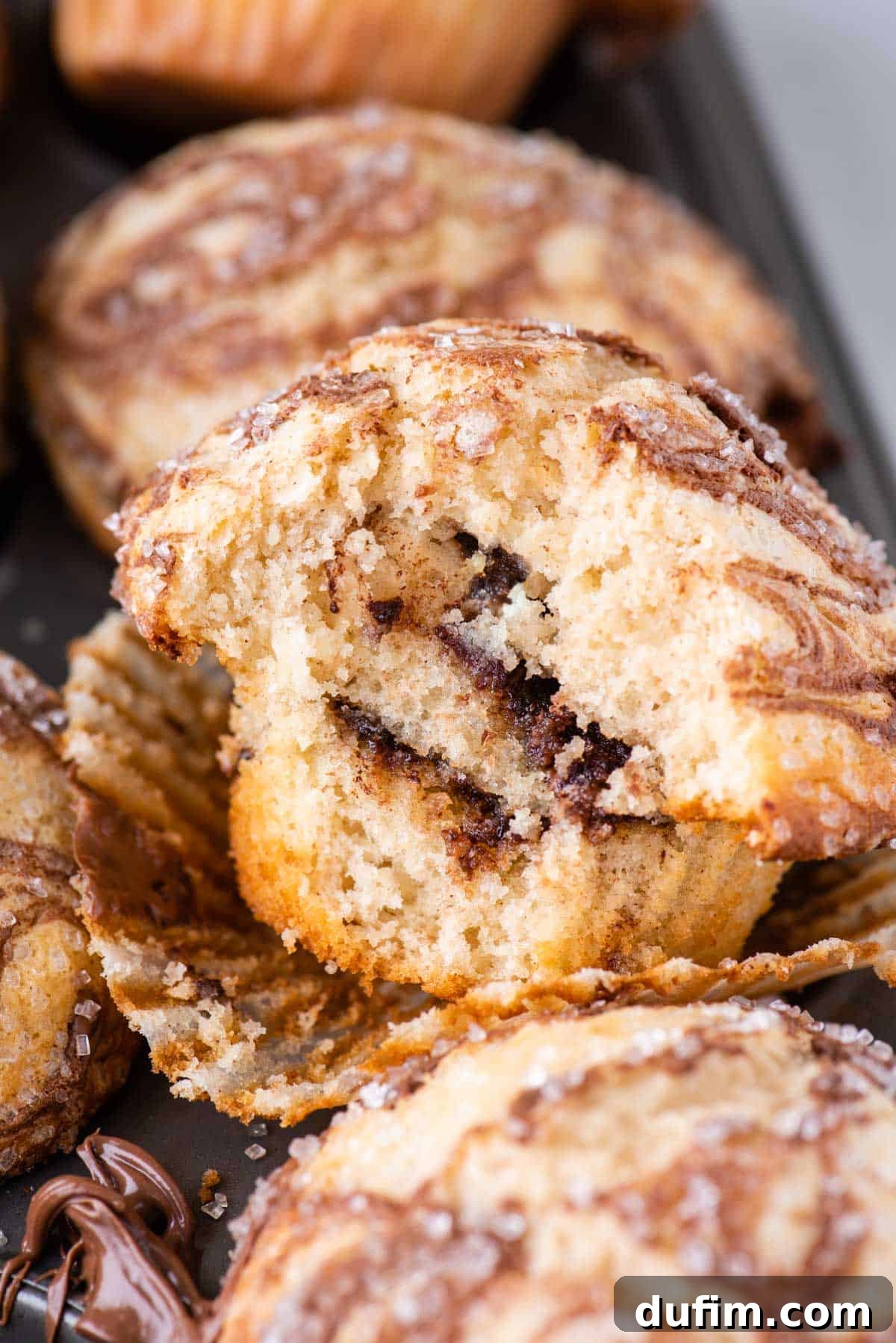 A Nutella swirl muffin with a bite taken out, clearly exposing the delicious layers of Nutella inside. It rests in its opened muffin liner, sitting on top of a muffin pan filled with more freshly baked muffins.