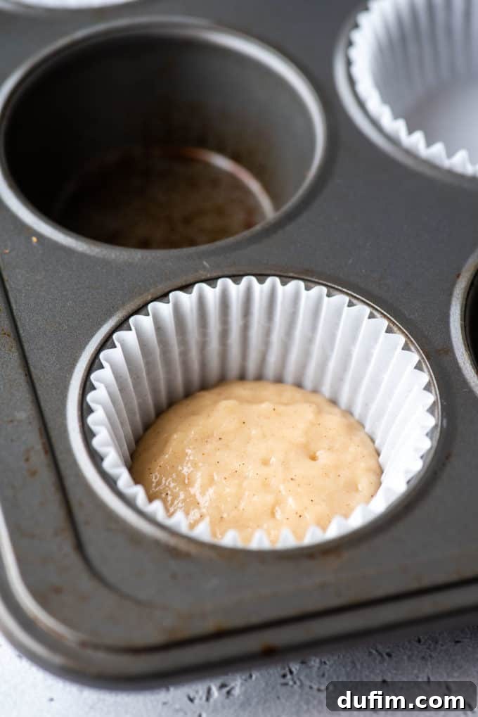 A muffin pan with a white muffin liner positioned in one of the corner spots, filled with a small amount of muffin batter at the bottom.