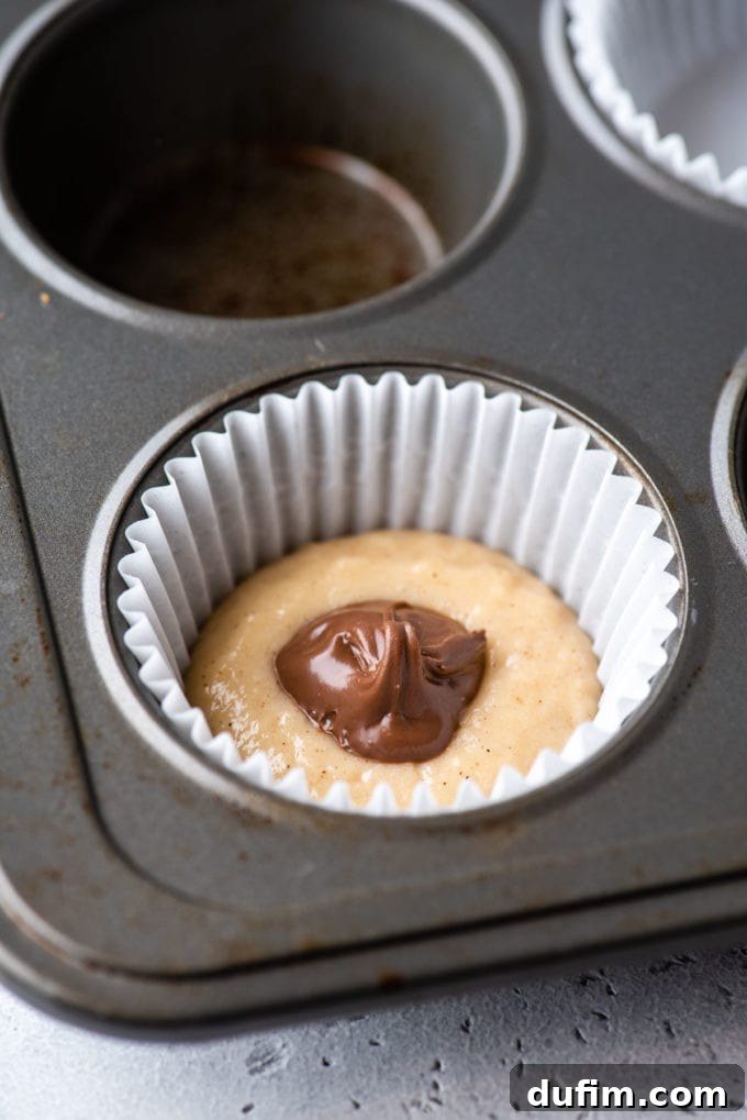 A muffin pan with a white muffin liner in a corner spot, containing muffin batter at the bottom and a generous dollop of Nutella placed directly on top of the batter.
